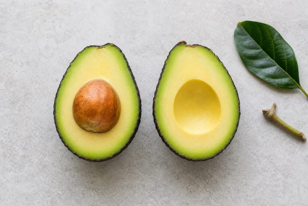 Ripe avocado cut in half showing the dark skin, pale green flesh and large brown pit, with a leaf beside it