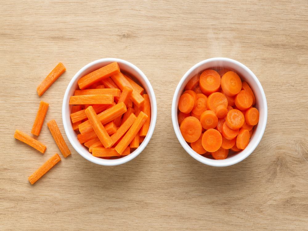 Raw carrot sticks beside lightly steamed carrot rounds in two identical bowls